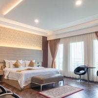 Habitación de hotel con cama, banca, silla, mesita pequeña y ventana con cortinas.