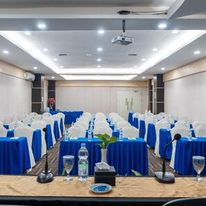 A conference room with rows of tables, each with a blue tablecloth, white chair covers, water bottles, and a microphone. - Muara Hotel and Mall Ternate (Ternate)