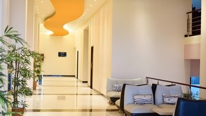 Un vestíbulo de hotel moderno con suelo brillante, una lámpara de techo curva, zona de estar con sofás y plantas en macetas.
