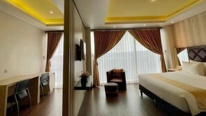 Habitación de hotel con cama, escritorio, silla, sofá y un amplio ventanal con cortinas.