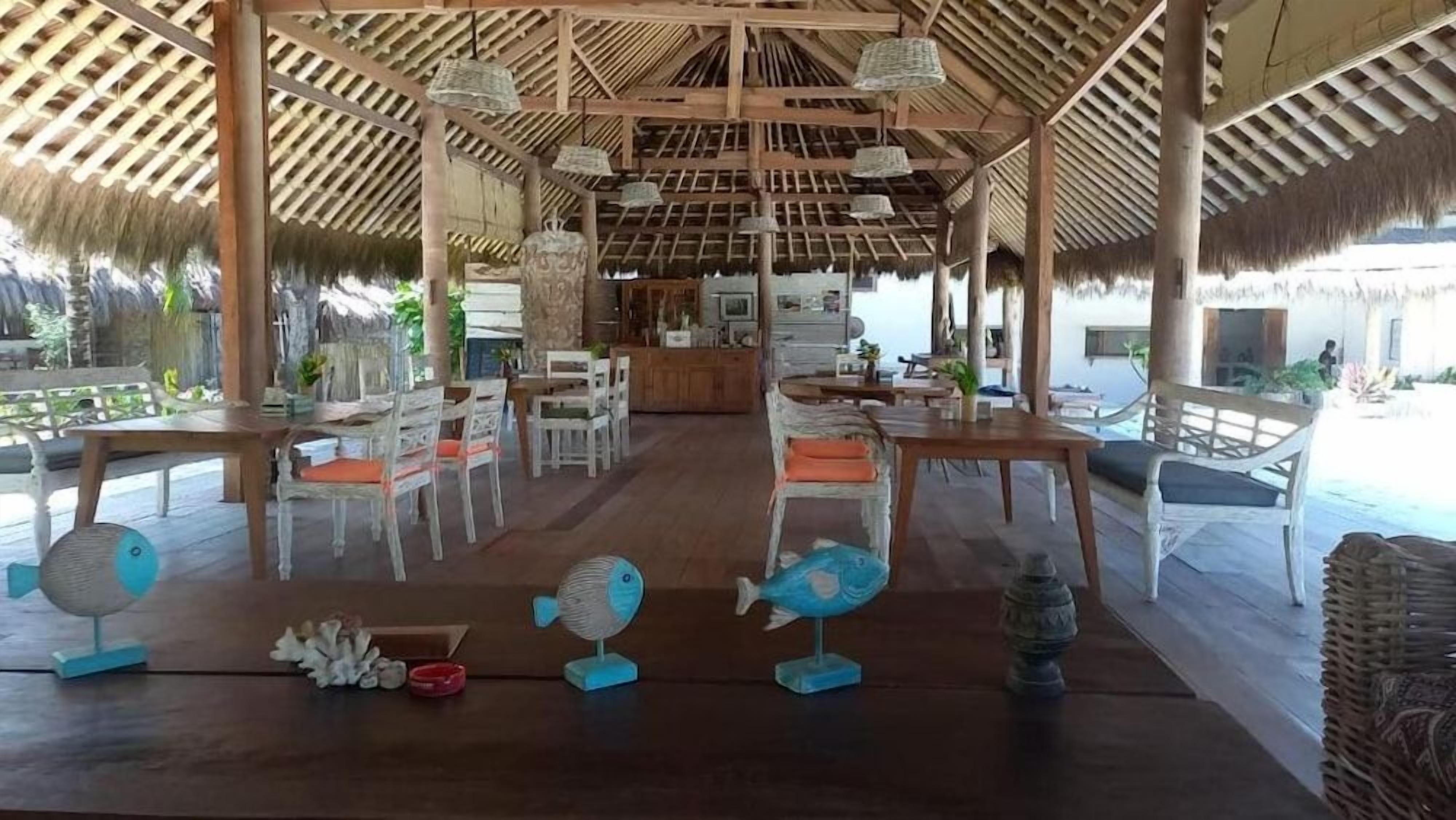 An open-air dining area with wooden tables and chairs, a thatched roof, and decorative fish sculptures.