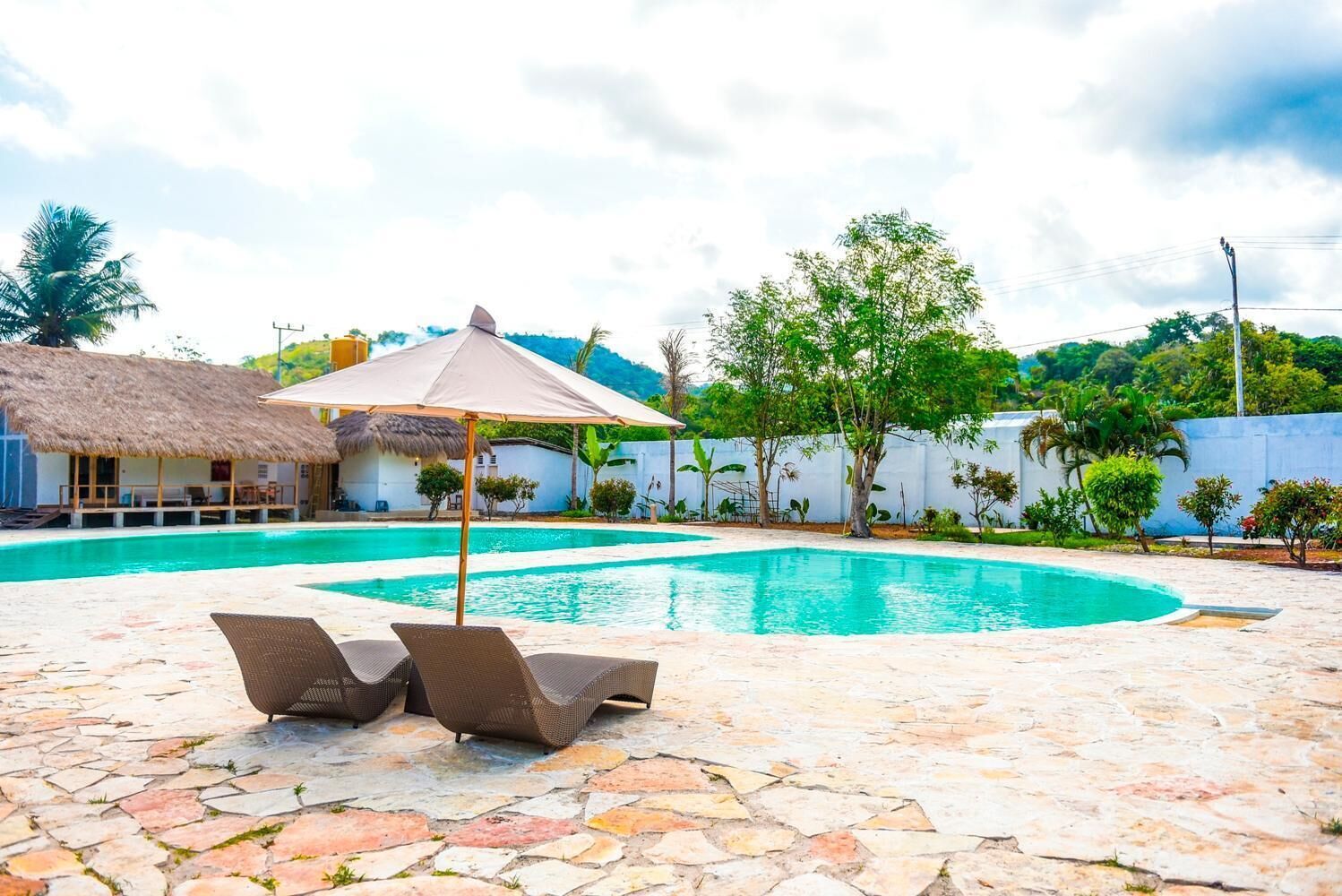 A swimming pool with lounge chairs and a shaded umbrella, surrounded by a stone patio and a thatched-roof structure.