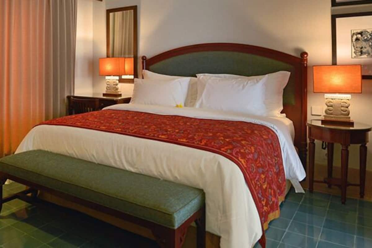 A hotel room with a bed, a chair, a bench, two bedside tables with lamps, and a curtain.