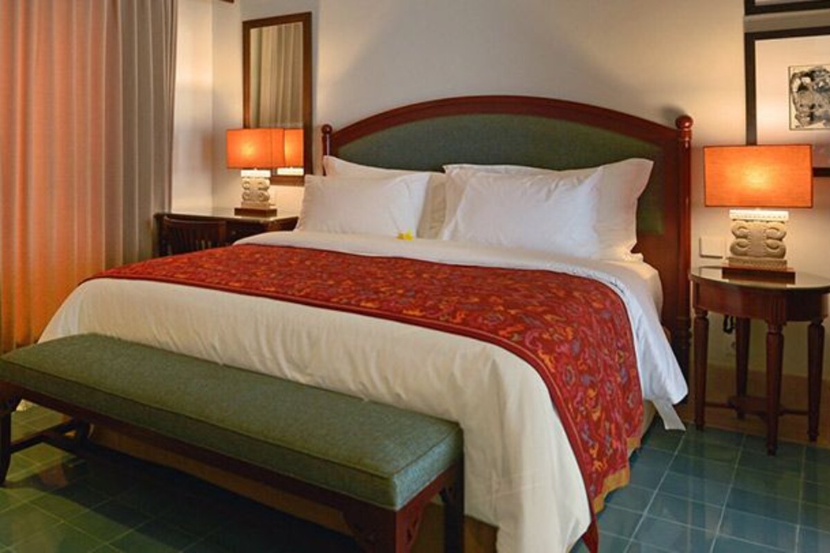 A hotel room with a bed, a chair, a bench, two bedside tables with lamps, and a curtain.