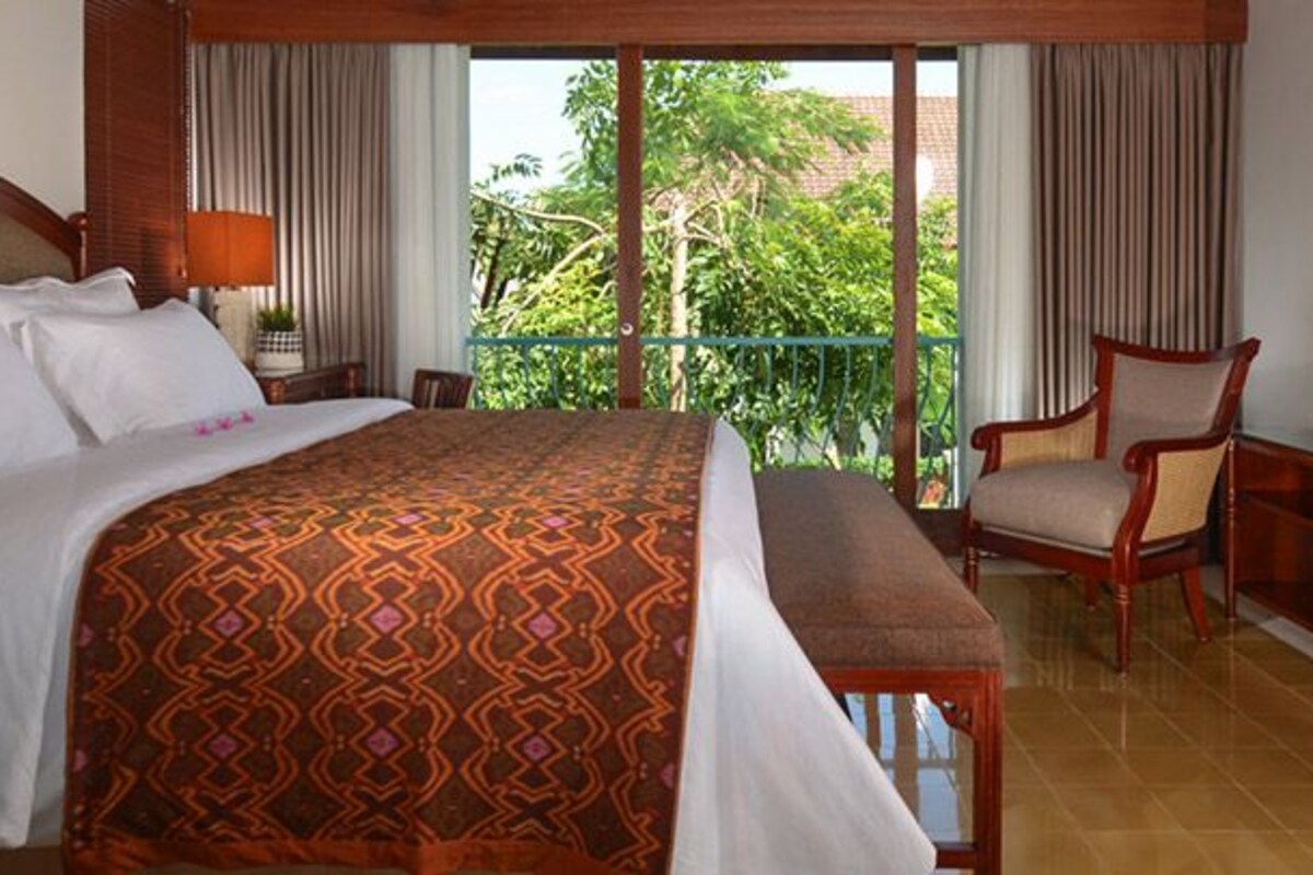 A hotel room with a large bed, a chair, a table with a lamp, and a view of greenery outside.