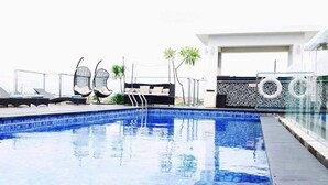 A swimming pool with lounge chairs and a sunbed, surrounded by a white building with a patterned fence. - Hotel ZIA Bali - Kuta (Kuta)