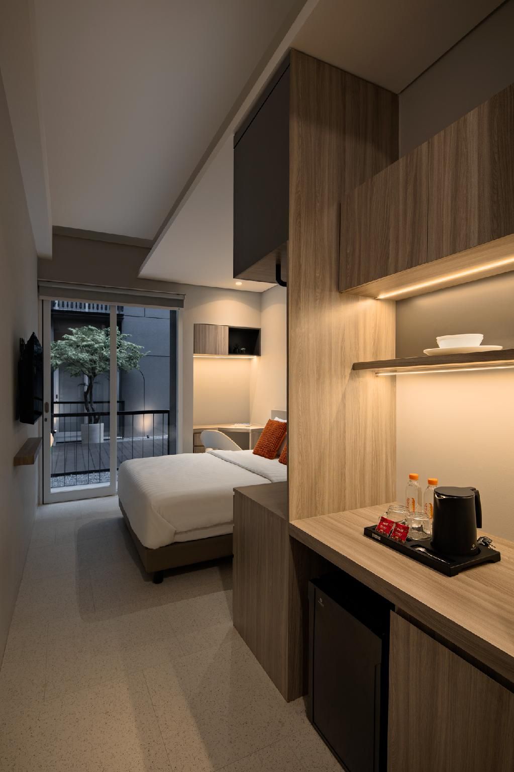 A modern hotel room with a bed, a small kitchenette, and a view of a tree through a sliding door.