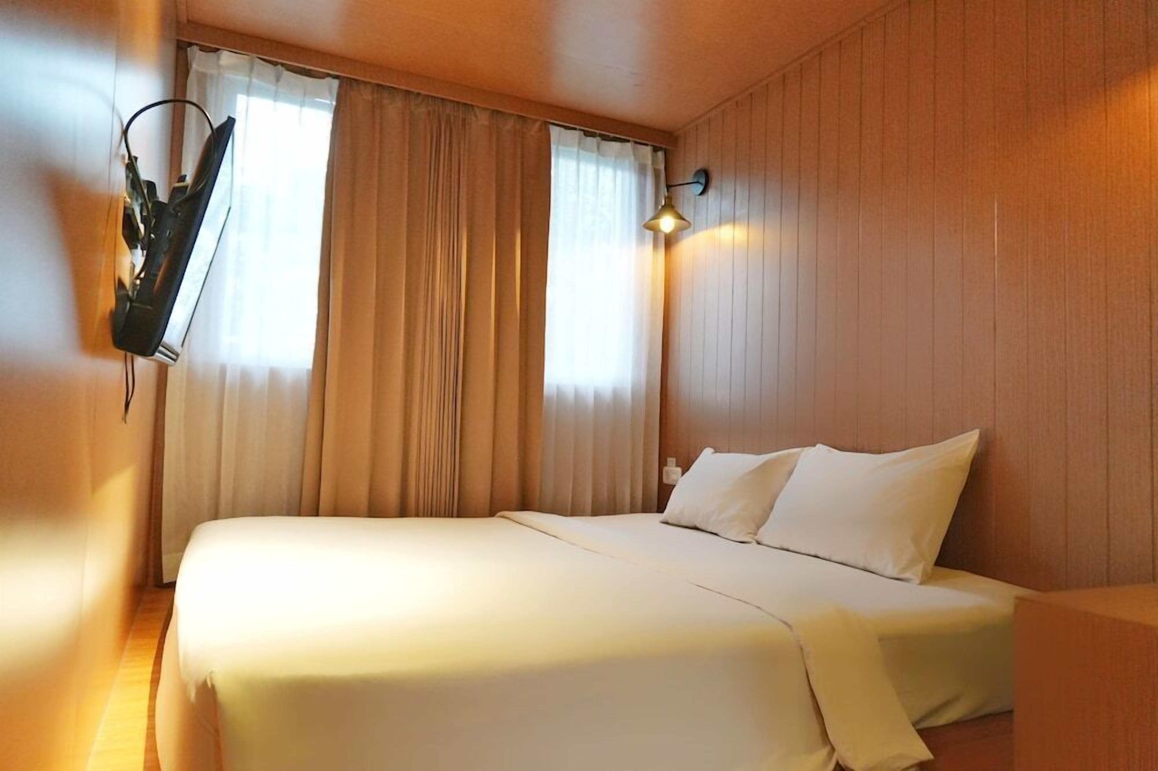 A hotel room with a bed, curtains, a wall-mounted lamp, and a flat-screen TV.