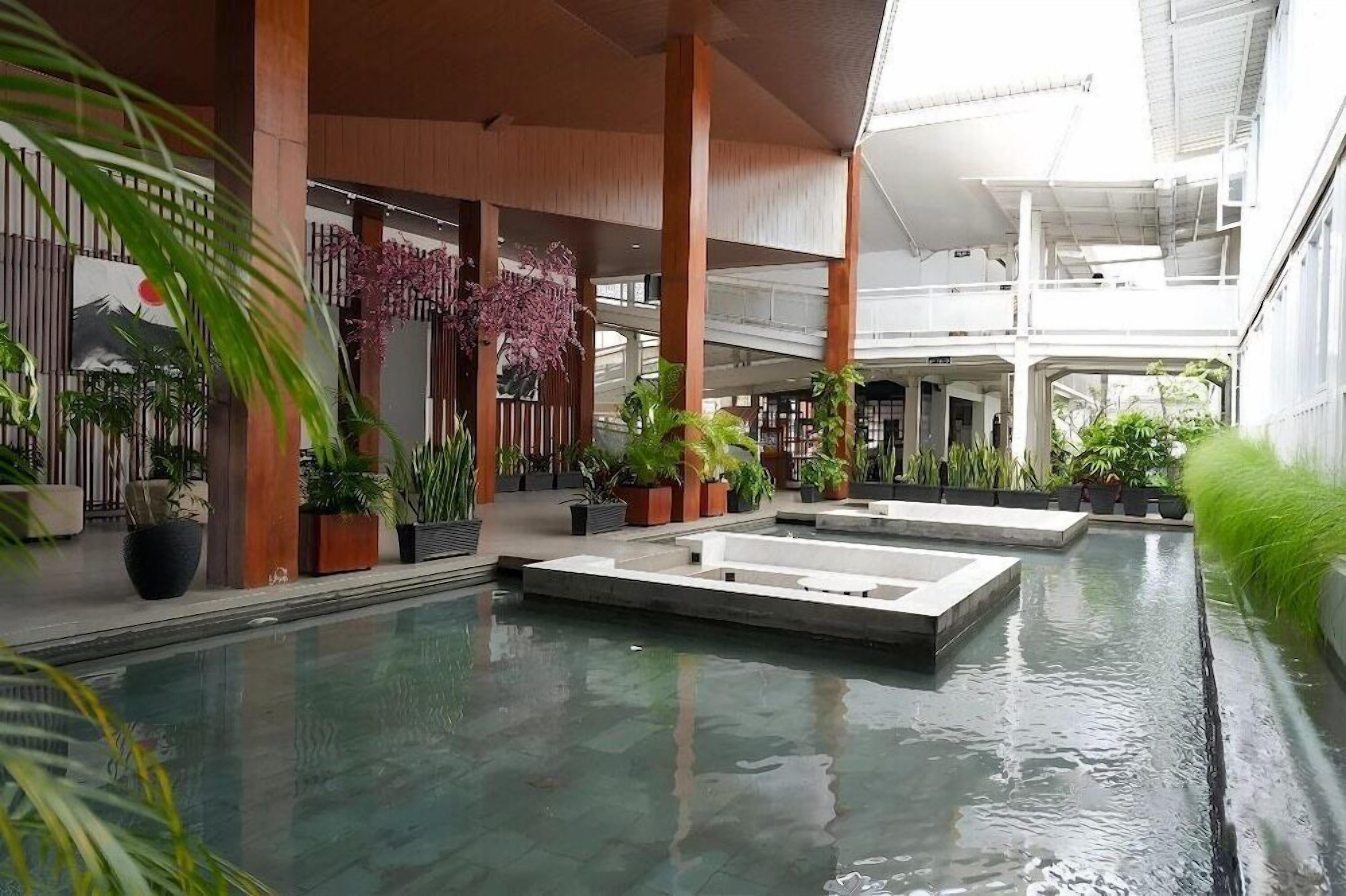 An indoor swimming pool area with multiple lanes, surrounded by potted plants and wooden pillars.