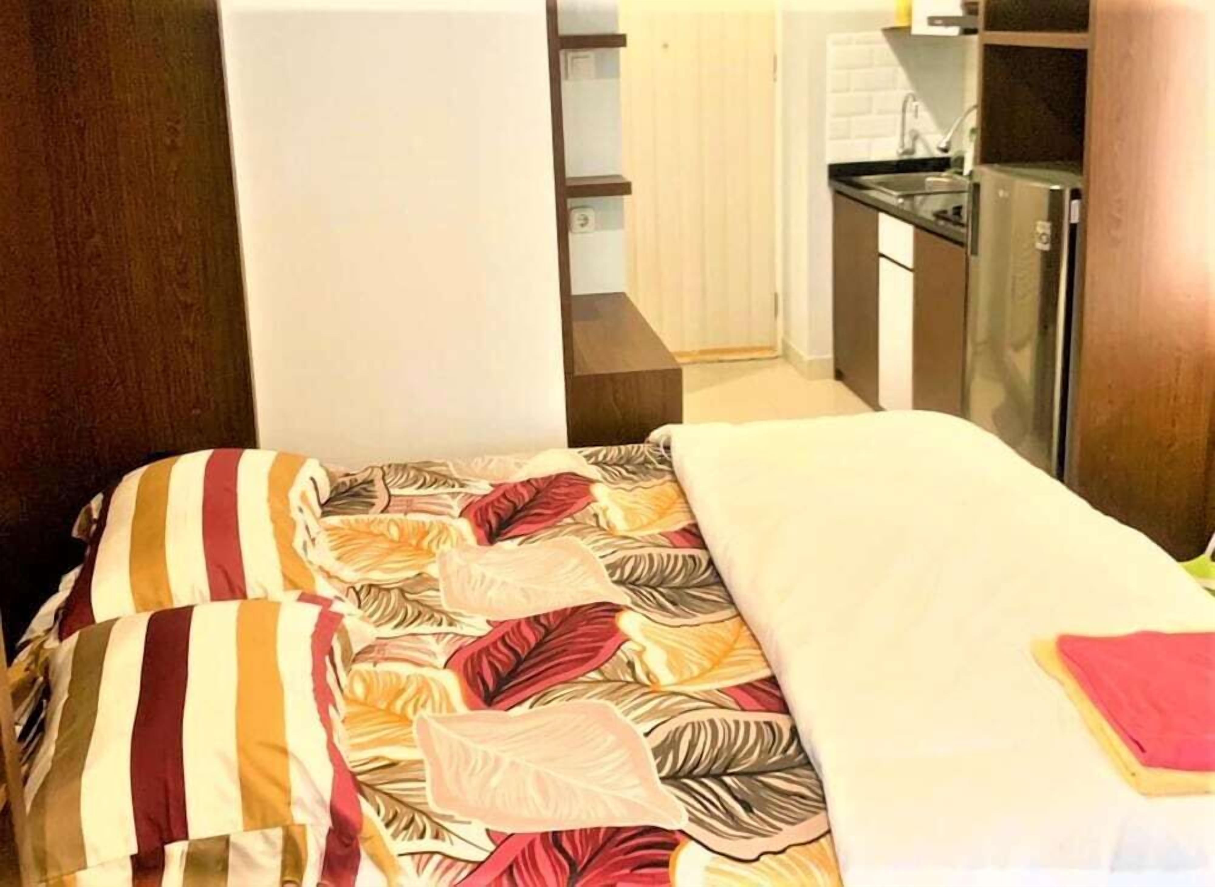 A bedroom with a bed, pillows, and a bedside table. There is a kitchenette with a sink and a stove in the background.
