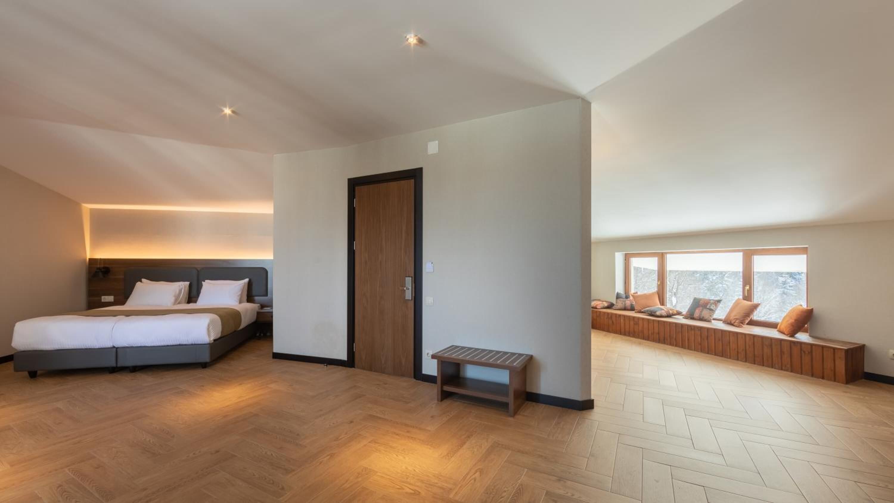 A modern hotel room with a large bed, a bench, and a seating area by the window.