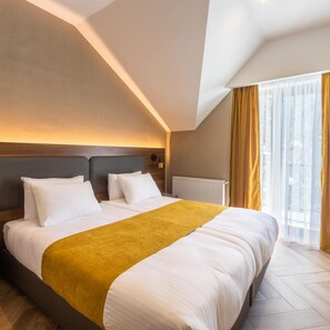 Een hotelkamer met een groot bed, een hoofdeinde, een nachtkastje, een raam met gordijnen en een radiator.