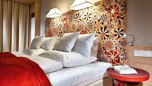 Une chambre d’hôtel avec un lit, une couverture rouge, une table ronde sur laquelle se trouvent un livre et des lunettes, et une tête de lit ornée d’un motif floral.