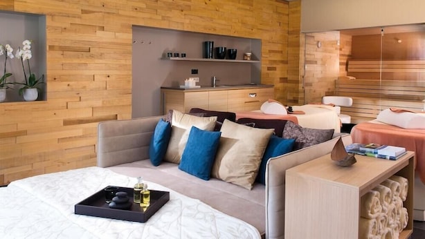 Une chambre d’hôtel moderne avec un mur en bois, un canapé avec des coussins, une table de chevet sur laquelle se trouve un plateau de produits de toilette, et un petit coin salon avec une chaise et une table.