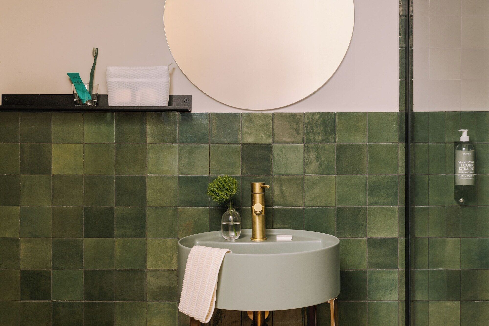 A bathroom with a round sink, green tiles, a mirror, and toiletries.