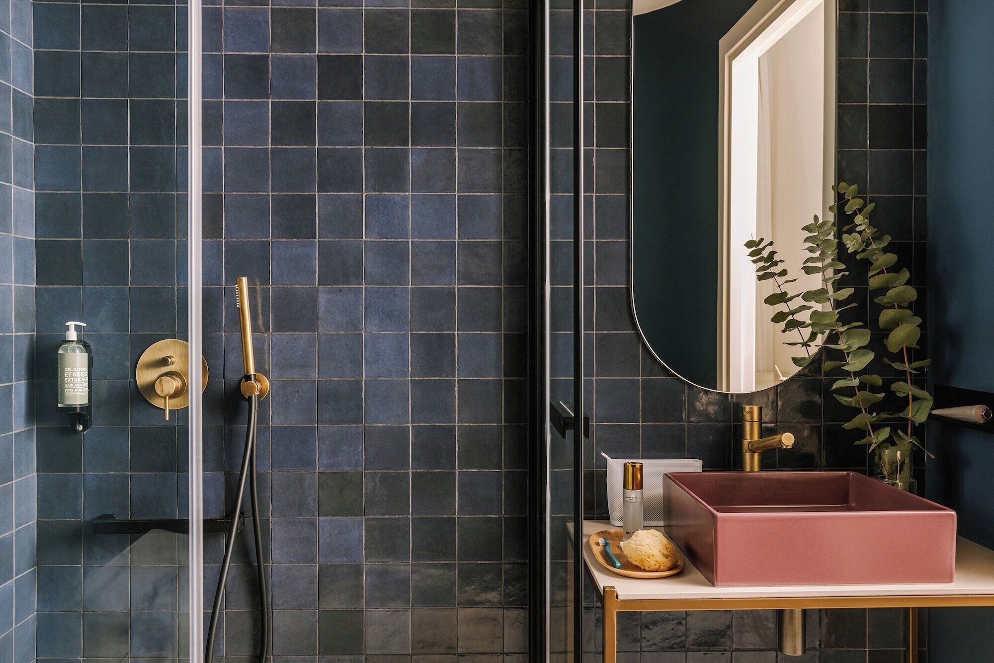 Casa de banho com azulejos azul escuro, um chuveiro dourado, um espelho e uma pia rosa.