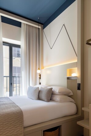 A hotel room with a bed, a nightstand, a lamp, and a view of a building through a window. - Ikonik Jean Medecin (Nice)