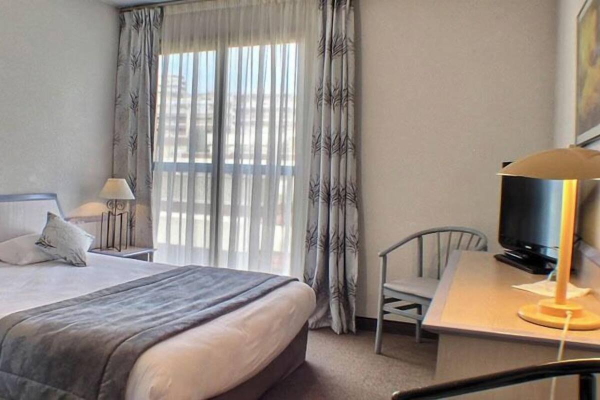 Une chambre d’hôtel comprenant un lit, des tables de chevet, un bureau avec une chaise, une télévision et une fenêtre avec des rideaux.