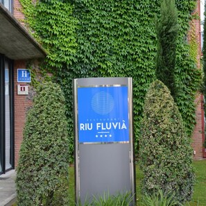 Skilt for Riu Fluvia restaurant med blå baggrund og hvid skrift, foran en bygning med murstensfacade og vægge dækket af slyngplanter.