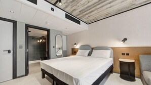 Una habitación de hotel moderna con una cama grande, mesitas de noche, un espejo y un pequeño rincón de descanso.