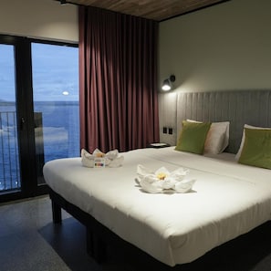 Quarto de hotel com cama, mesa de cabeceira, luminária, vista para o mar e cortina.