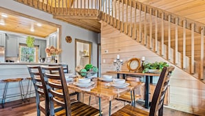Un coin repas avec une table en bois dressée pour six personnes, entourée de chaises en bois. Un escalier en colimaçon avec des rampes en bois est visible au fond.