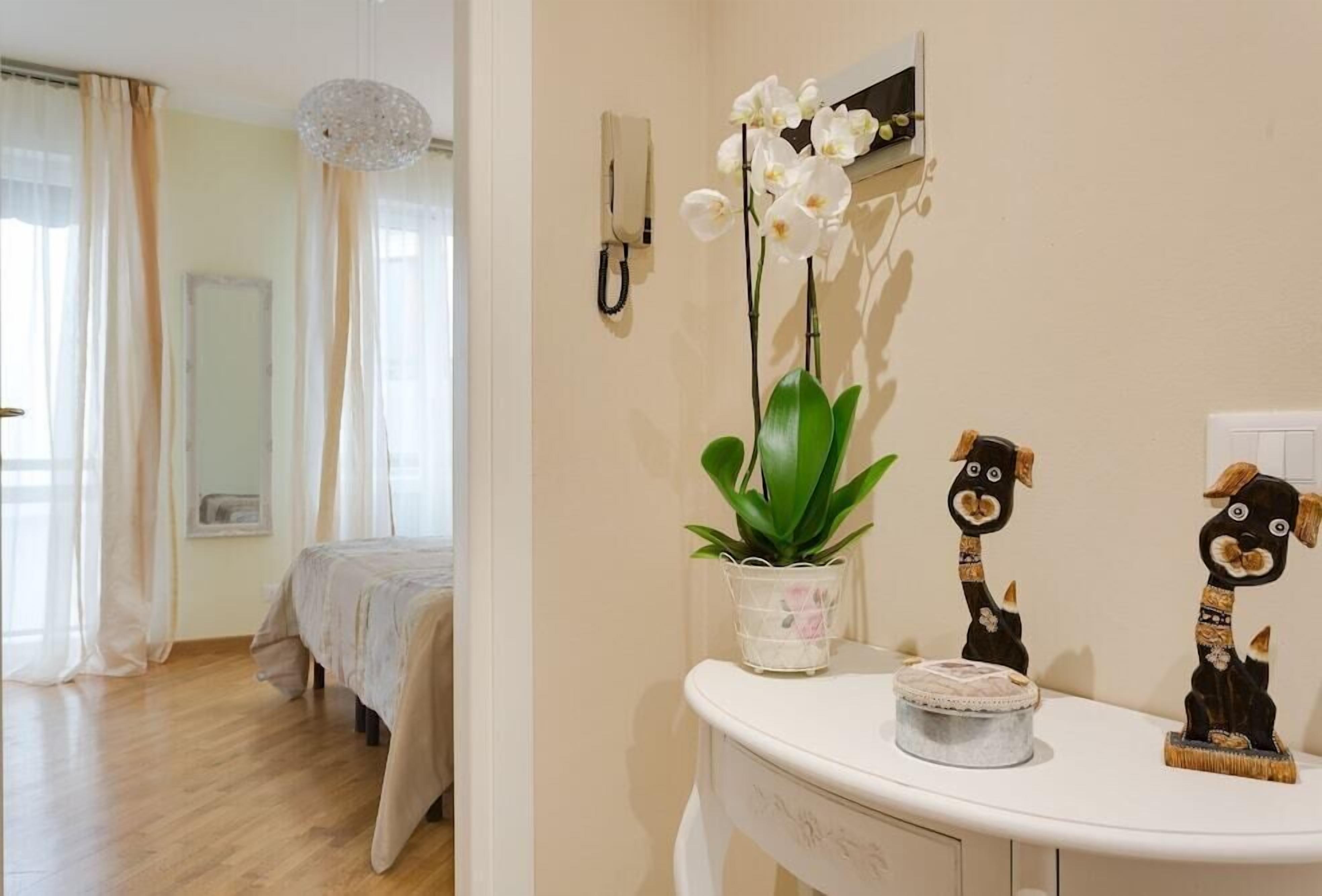 A hotel room with a bed, a small table with a plant and decorative items, a wall-mounted phone, and a window with curtains.