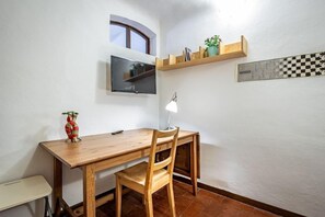 A small room with a wooden desk, a chair, a television mounted on the wall, a small shelf with books and a plant, and a window with a view. - Flospirit - Viola (Florence)