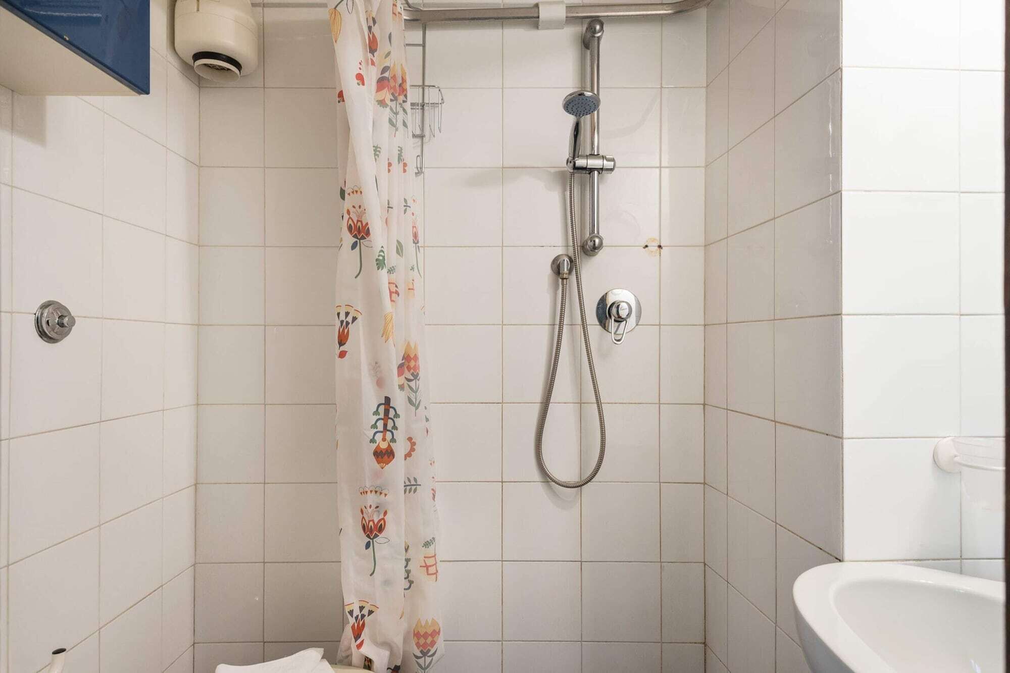 Een badkamer met witte tegels, een douche met handdouche, een toilet en een gordijn met bloemenmotieven.