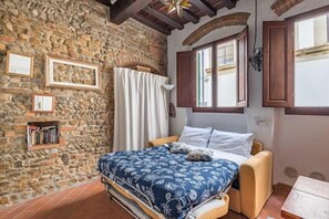 A bedroom with a stone wall, a bed with a floral bedspread, a window with wooden shutters, and a bookshelf. - Mini Studio in the Heart of Florence (Florence)