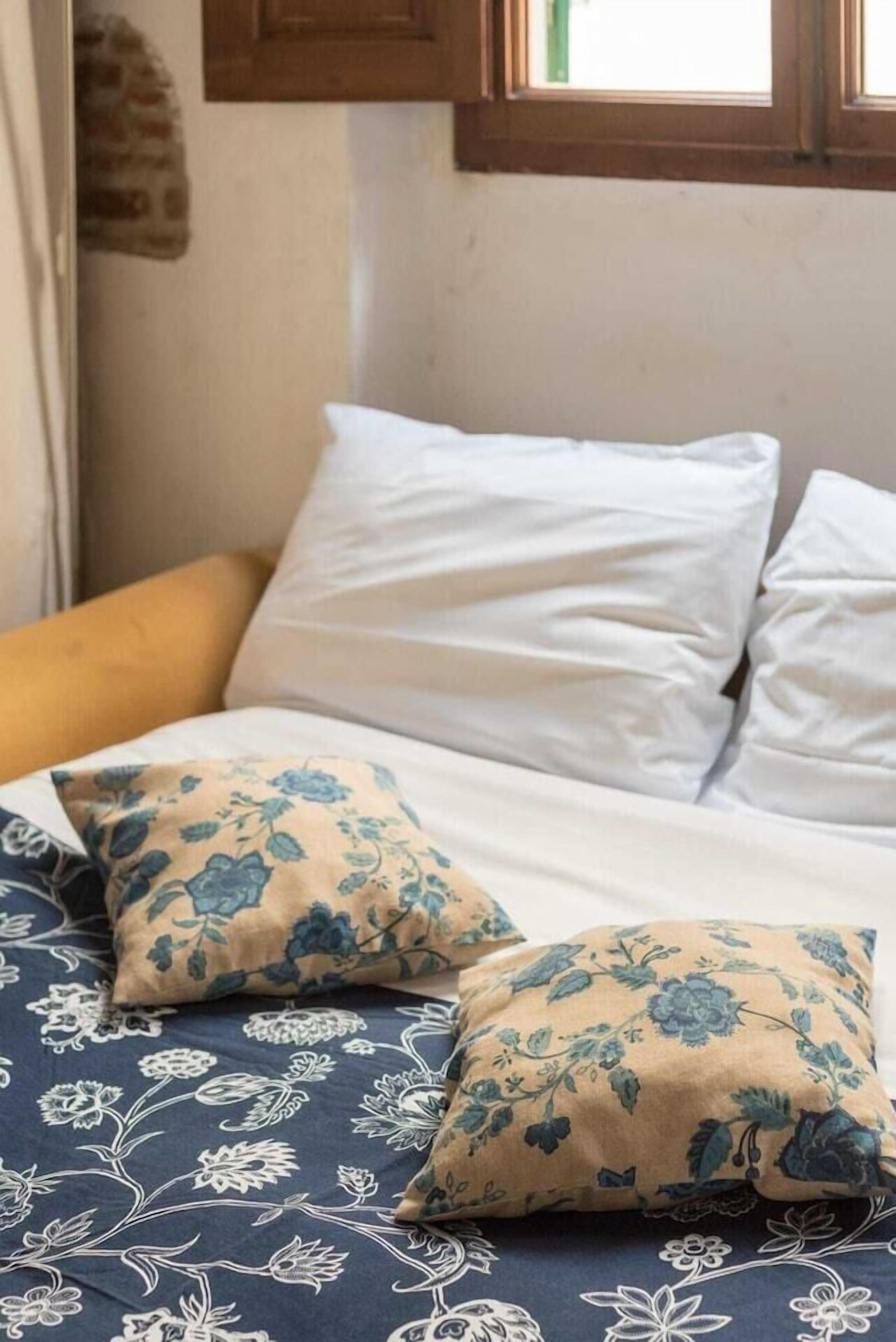 A bed with white and blue floral bedding, a yellow pillow, and a window with wooden frames.
