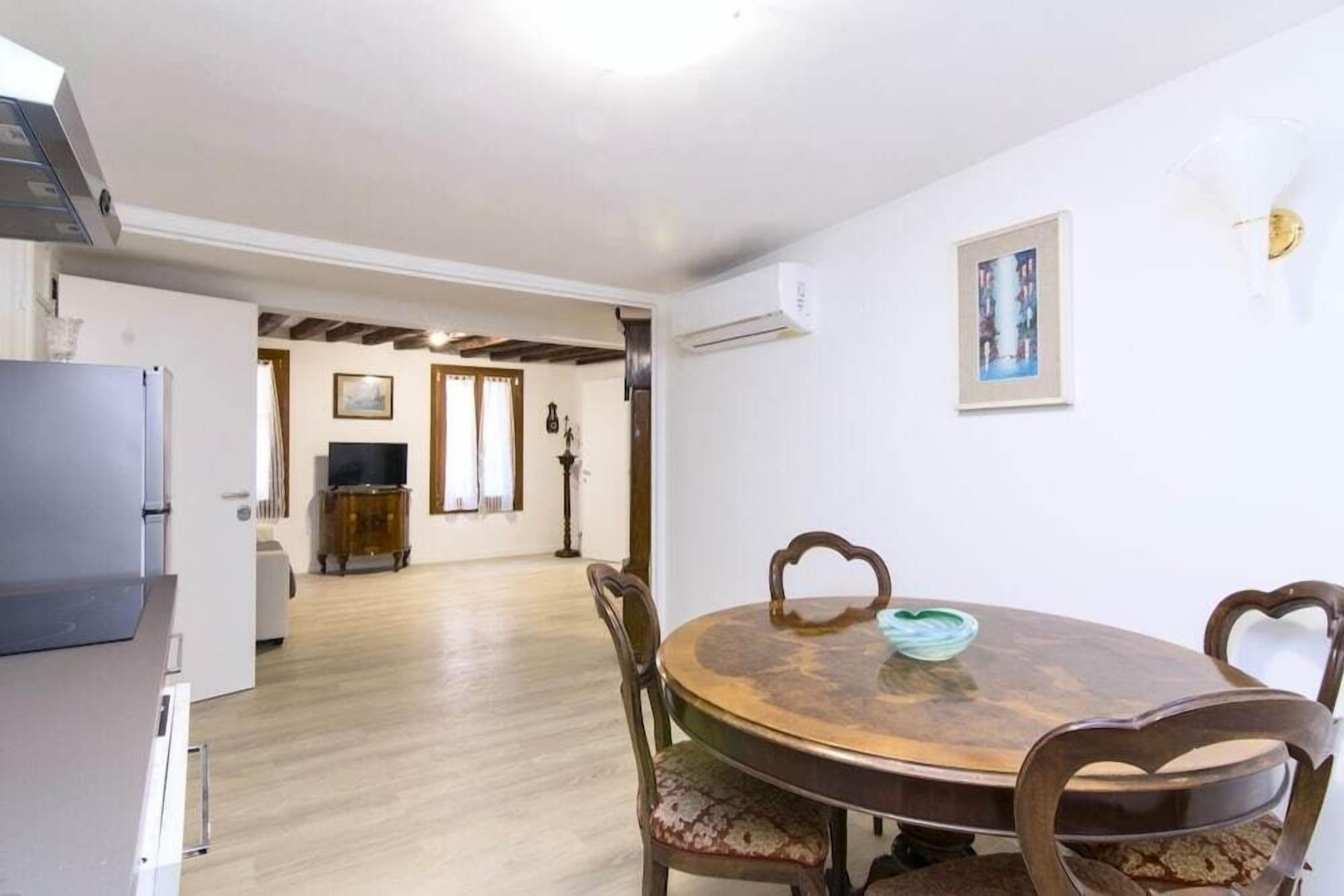 A dining area with a round wooden table and chairs, a television in the background, and a hallway leading to other rooms.