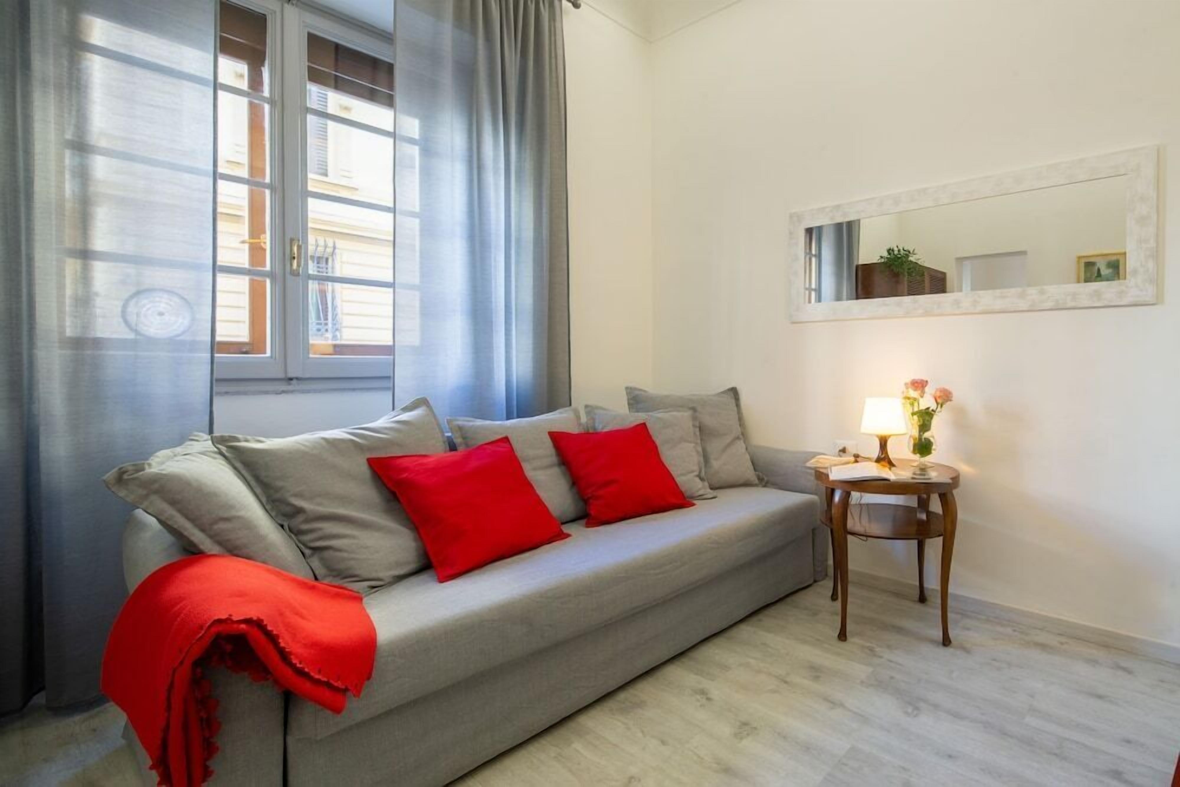 A living room with a grey sofa, red cushions, a small wooden side table with a lamp, and a mirror on the wall.