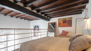 A bedroom with a large bed, wooden beams, a window with a view, a painting, and a lamp.