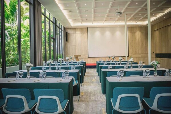 Un salón de conferencias con mesas dispuestas para una reunión, ventanas amplias y una pantalla de proyector.