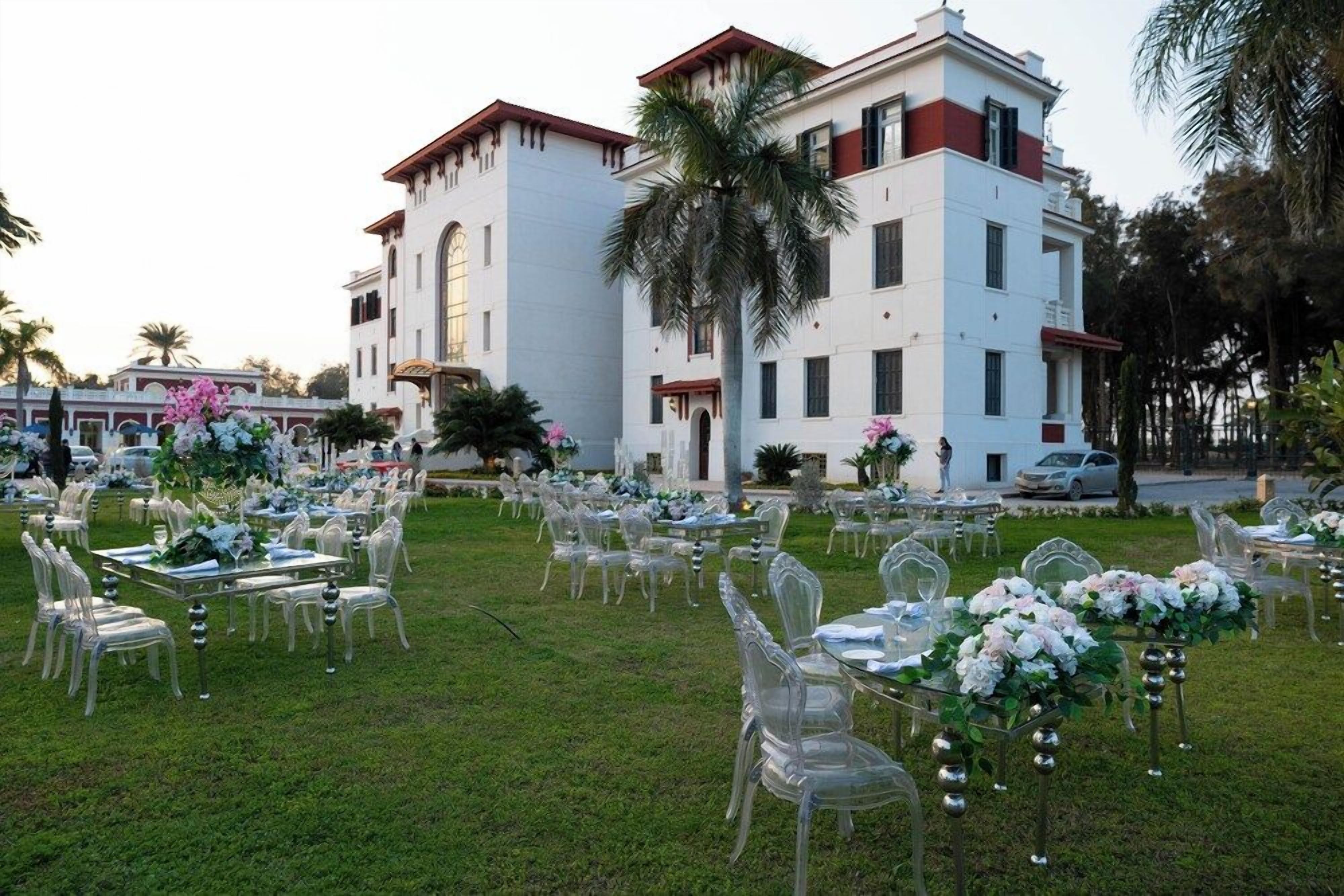 An outdoor wedding venue with tables and chairs arranged on a lawn, a large white building in the background, and palm trees.