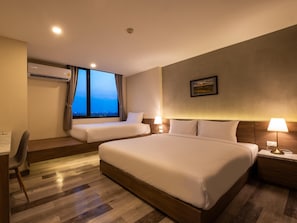 A hotel room with two beds, a desk, a chair, a window with curtains, a wall-mounted air conditioner, and a nightstand with a lamp. - B2 Korat Premier Hotel (Nakhon Ratchasima)