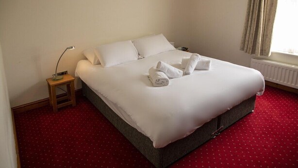 A neatly made bed with white linens and towels, a small bedside table with a lamp, and a red carpeted floor. - The Knighton Hotel by Payman Club (Knighton)