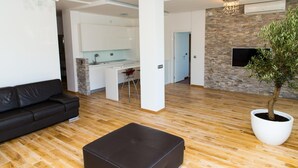 A modern living room with a black leather sofa, a wooden floor, a large potted plant, and a kitchen area in the background. - Hotel Ponta Nova (Bečići)