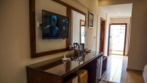 A hotel room with a TV mounted on the wall, a wooden vanity with a mirror, a small seating area, and a view of the outside.