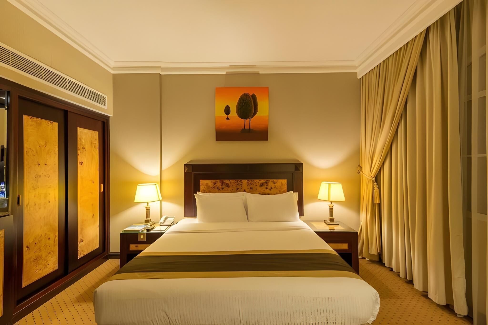 A hotel room with a large bed, two bedside tables with lamps, a painting on the wall, and a window with curtains.