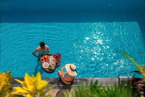 Cena à beira da piscina com uma pessoa sentada na borda, uma bandeja com flores e uma bebida, e outra pessoa em pé na água.