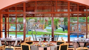 A restaurant with a view of a landscaped garden featuring a zebra and a hippo. Tables are set with white tablecloths and glassware.