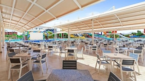 A covered outdoor dining area with multiple tables and chairs, a pool, and a water park in the background.