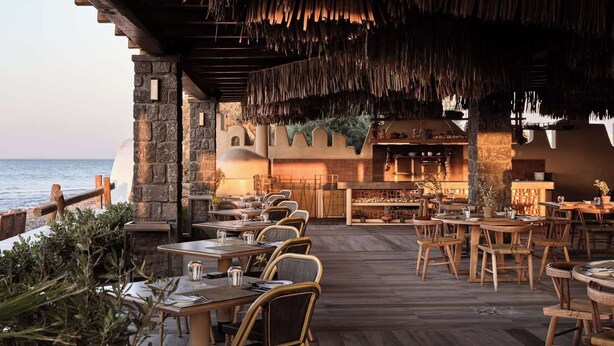 Un restaurant en plein air avec des tables et des chaises en bois, un mur en pierre et une vue sur la mer.