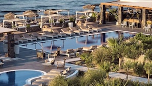 Un espace piscine aménagé dans un complexe hôtelier, avec des chaises longues et des parasols, entouré de palmiers et offrant une vue sur l’océan.