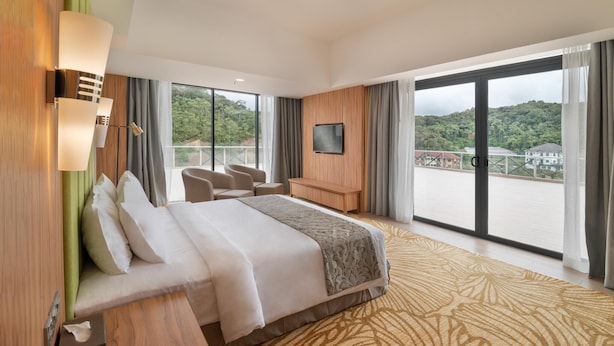 Habitació d'hotel amb un llit gran, escriptori, televisió i un balcó amb vista a vegetació.