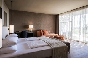 Et hotelværelse med en stor seng, et skrivebord med lampe og udsigt til det fri gennem lette gardiner.