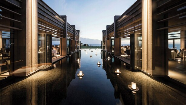 Un couloir d’hôtel moderne avec un revêtement de sol réfléchissant, des portes en verre et une vue sur la mer.