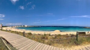 A beach with a wooden boardwalk leading to the shore, clear turquoise waters, and a few buildings in the background. - Sa Pergola (Formentera)